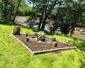 A campsite with a fire pit surrounded by logs and a barrel, set in a grassy area near a river.
