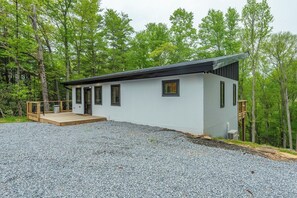 A modern single-story building with a gravel driveway, a wooden deck, and a forested backdrop.