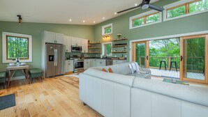 Eine moderne Küche mit weißen Schränken, einem Edelstahlkühlschrank und einer Mikrowelle. Es gibt einen Essbereich mit Tisch und Stühlen sowie ein großes Fenster mit Blick auf eine Terrasse.