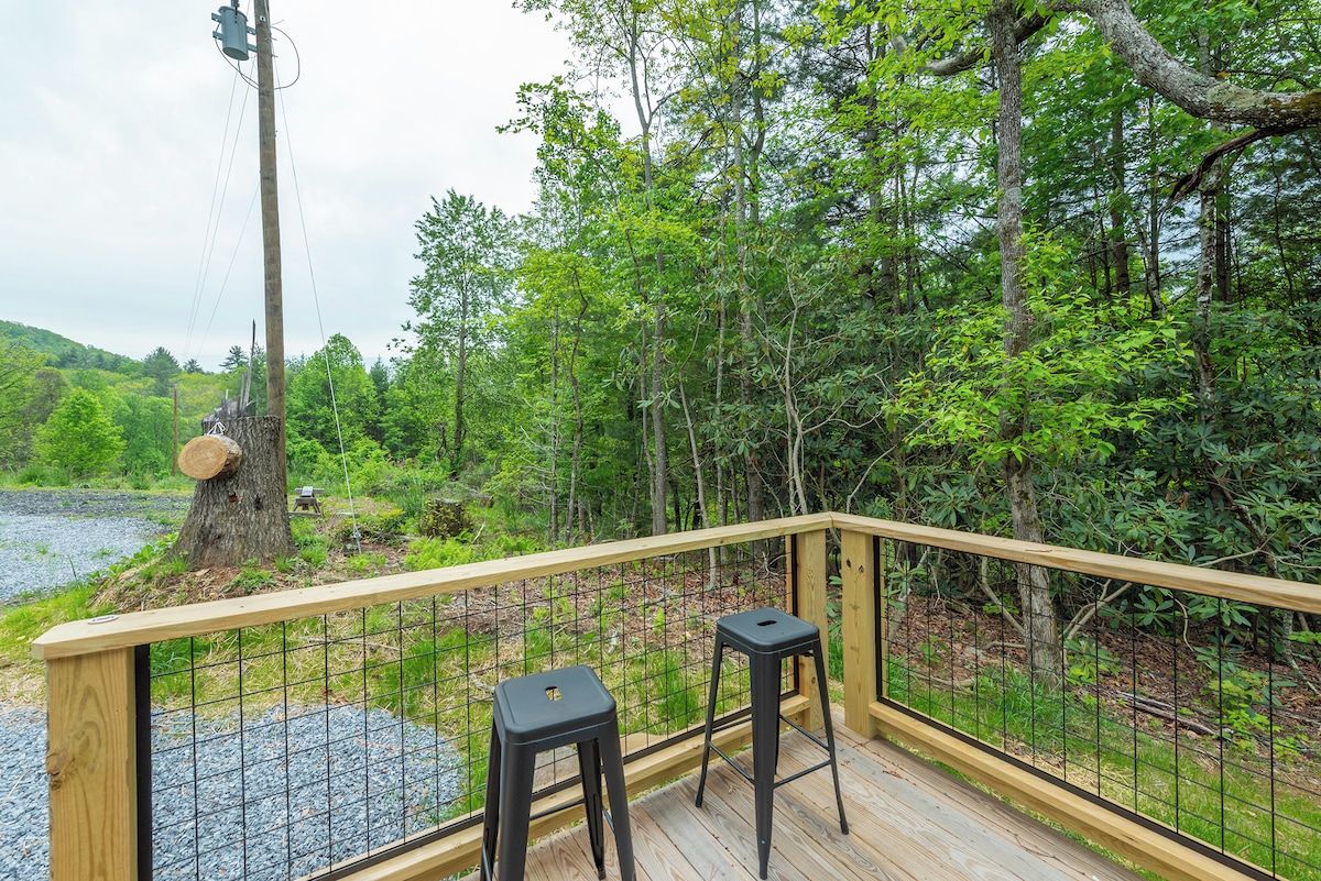 A wooden deck with two black stools, surrounded by a metal railing, overlooking a river and forested area.
