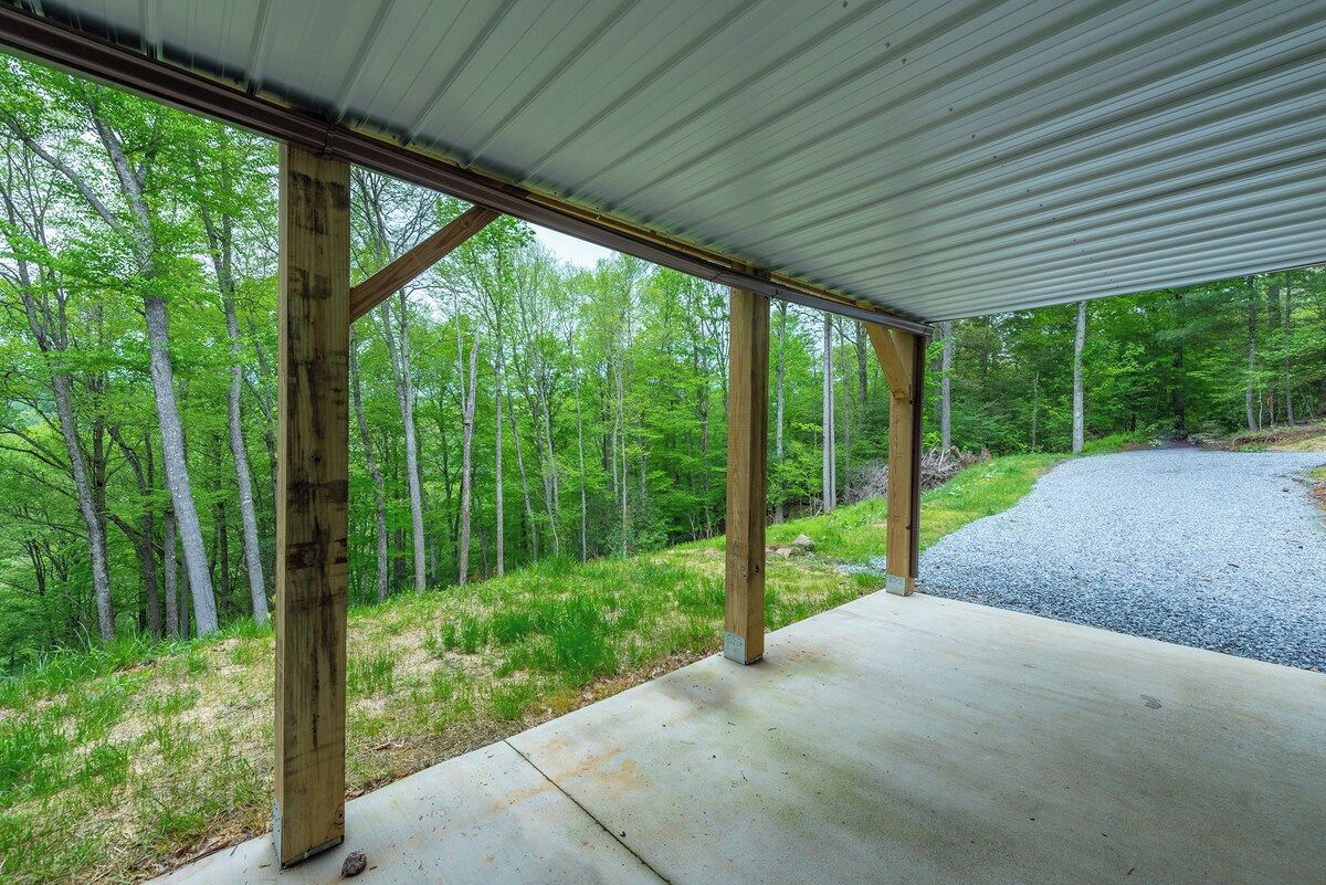 A covered outdoor area with a concrete floor and wooden posts, surrounded by a forested landscape.