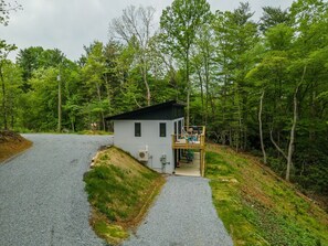 A small, single-story cabin with a porch, surrounded by dense forest.