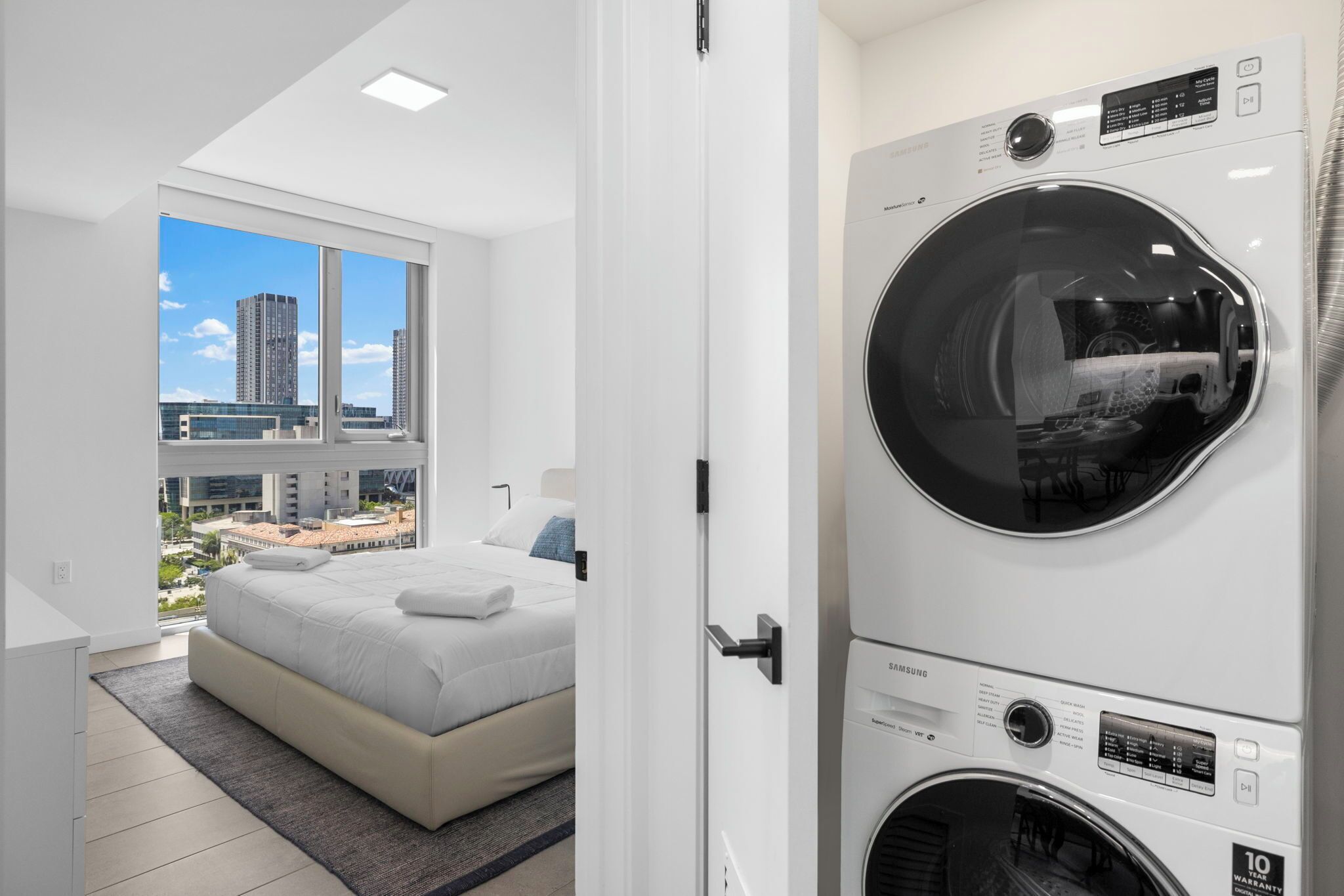 Habitación de hotel moderna con una cama grande, lavadora y vistas de la ciudad a través de amplias ventanas.