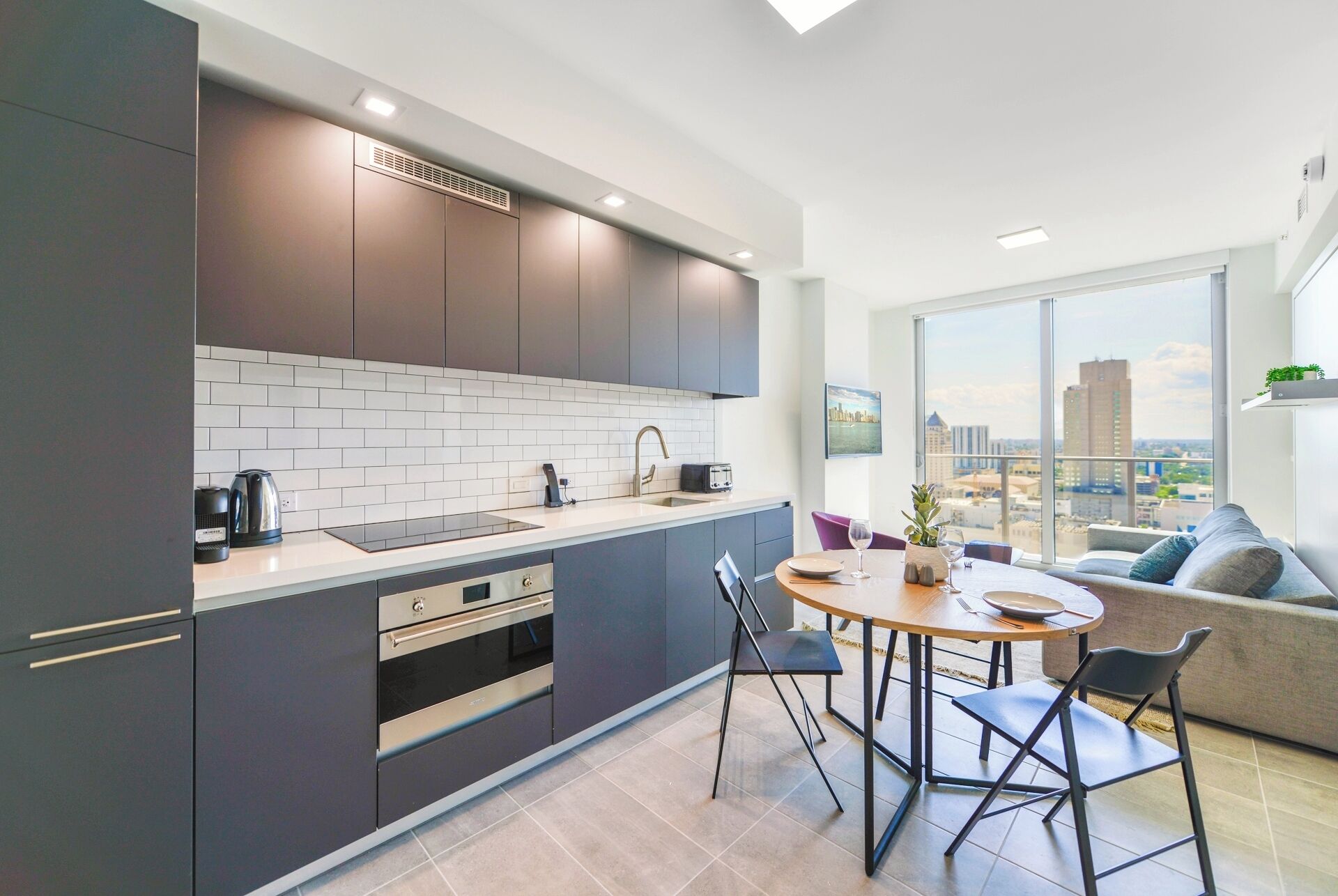 A modern kitchen with dark cabinets, a white tiled backsplash, and a city view through large windows.