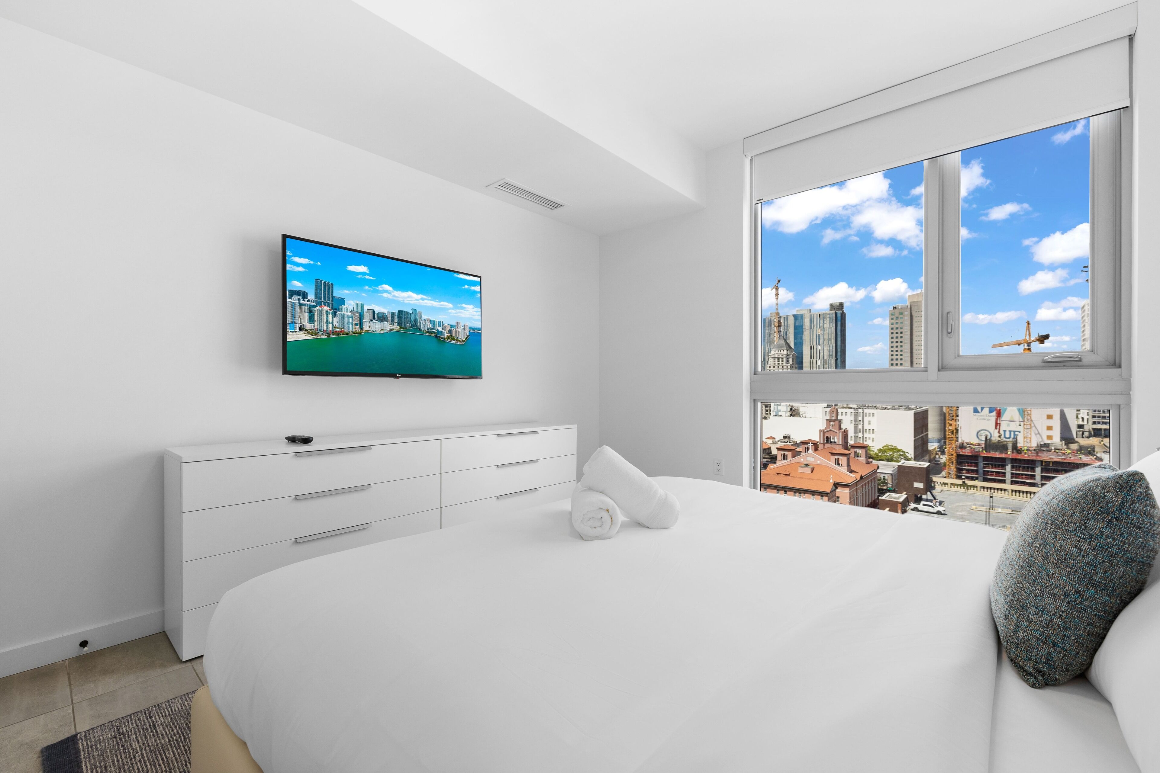 A modern hotel room with a large bed, a flat-screen TV on the wall, and a city view through the window.