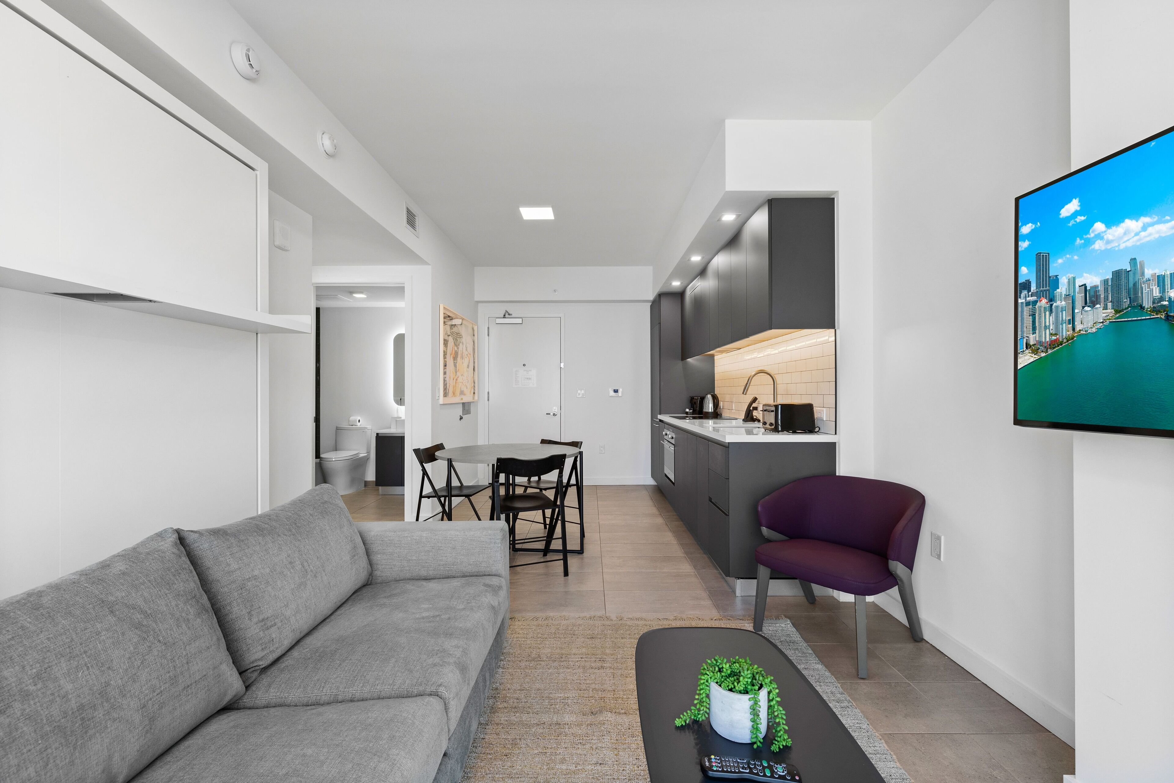 A modern living room with a grey sofa, a dining area with a table and chairs, a kitchen with dark cabinets, and a large TV on the wall.