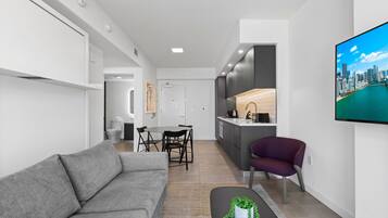 A modern living room with a grey sofa, a dining area with a table and chairs, a kitchen with dark cabinets, and a large TV on the wall.