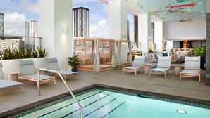 A modern indoor pool area with lounge chairs, a seating area with a wooden partition, and a view of tall buildings.