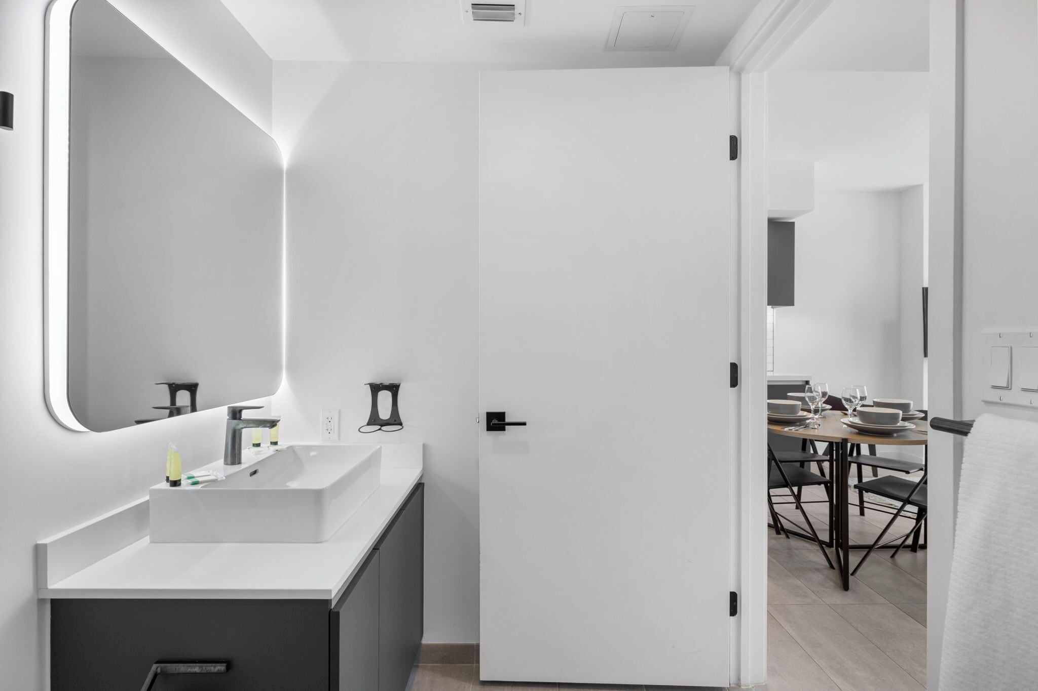 A modern bathroom with a white sink, mirror, and door leading to a dining area with a table set for two.