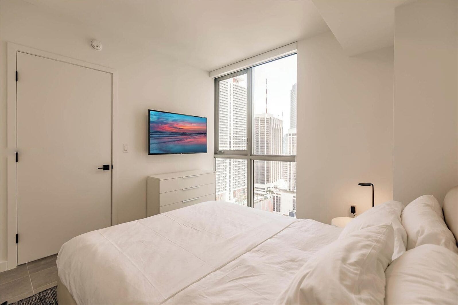 Une chambre avec un grand lit, une fenêtre donnant sur la ville, une table de chevet avec une lampe et un tableau accroché au mur.