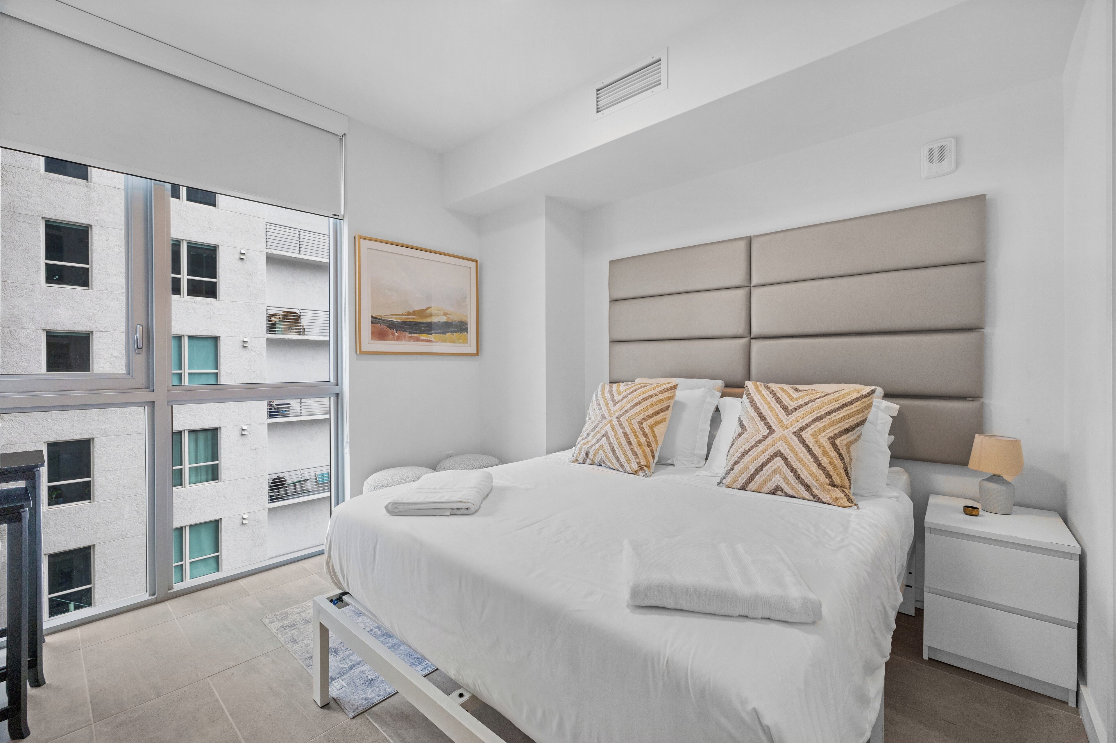 A modern bedroom with a large bed, a nightstand, a window with a view of a building, and a framed picture on the wall.