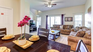 A living room with a sofa, a coffee table, and a TV. There is a dining table with plates and a flower vase.