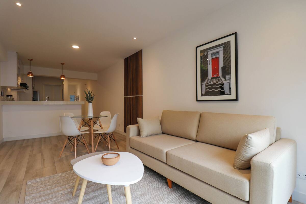 A modern living room with a beige sofa, a small white table, and a dining area with chairs and a glass table.