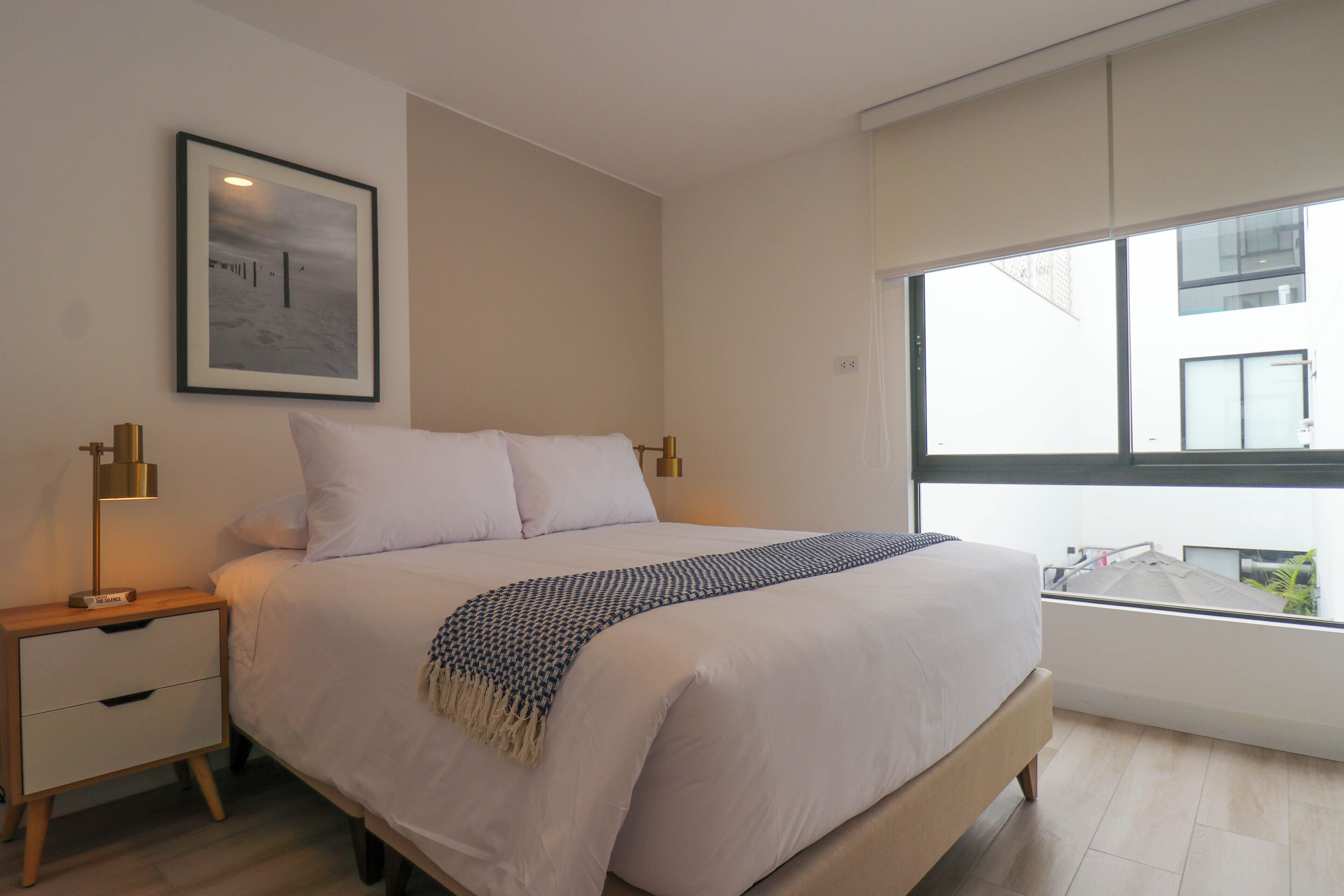 A modern bedroom with a large bed, bedside lamps, a framed picture on the wall, and a window with a view of buildings.