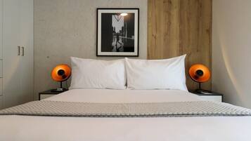 Cama bem arrumada com lençóis brancos, duas luminárias de cabeceira, um quadro na parede e um painel de madeira no lado direito.