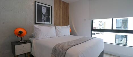 A modern bedroom with a large bed, a nightstand with a lamp, a framed picture on the wall, and a window with a city view.