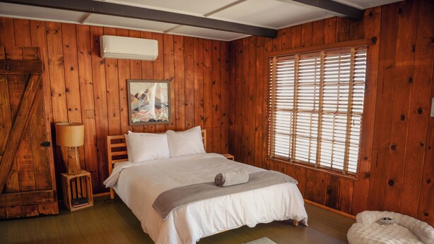 A wooden-paneled bedroom with a bed, a window with blinds, a lamp, and a small rug. - Room 6 • Retro Desert Escape  - Joshua Tree (Joshua Tree)