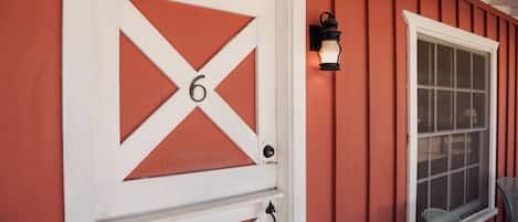A red barn-style building with a white door marked with the number 6, a window with a white frame, and a black wall-mounted lantern.