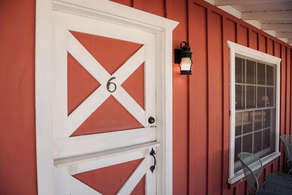 A red barn-style building with a white door marked with the number 6, a window with a white frame, and a black wall-mounted lantern. - Room 6 • Retro Desert Escape  - Joshua Tree (Joshua Tree)