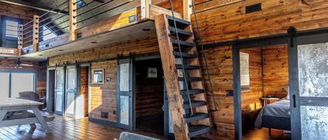 A spacious loft-style room with a wooden interior, a loft bed, a staircase, and a large window providing natural light.