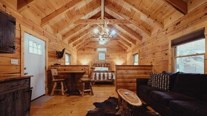 Interior de una cabaña de troncos con techo de madera, un candelabro, una cama con cabecera blanca, un sofá oscuro y una mesa de comedor con sillas.