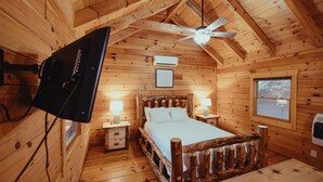 Habitación de cabaña de madera con cama, ventilador de techo, televisor montado y ventana con vistas.