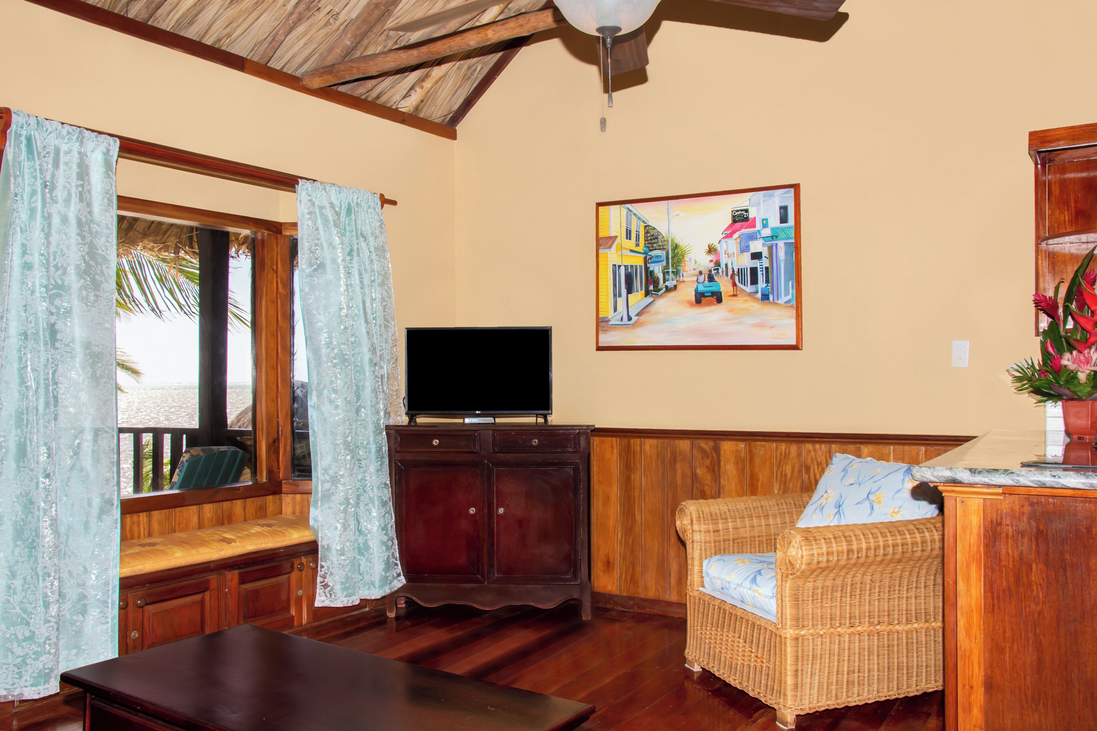 Una sala de estar con un banco de madera, un televisor sobre un mueble, un sofá de mimbre y un cuadro en la pared.