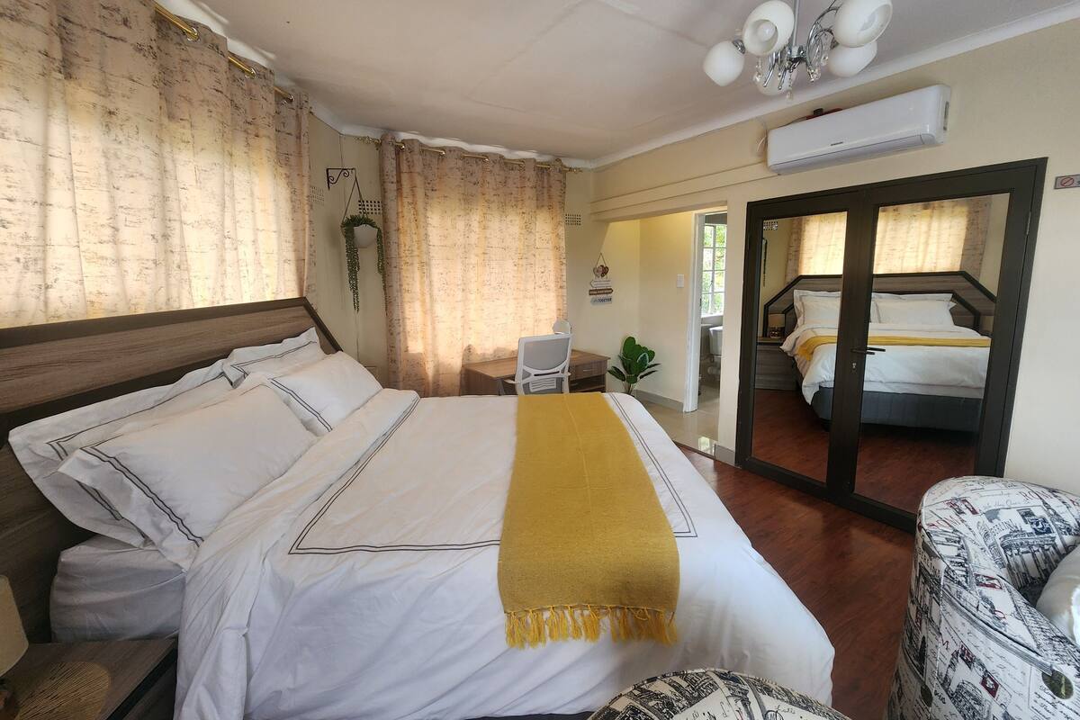 A hotel room with a bed, a desk, a chair, a mirror, and a door leading to another room.