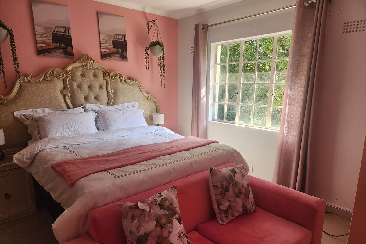 A bedroom with a bed, a nightstand, a window with curtains, and a pink sofa with floral cushions.