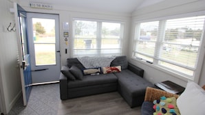 Una sala moderna con un sofá seccional gris, una mesita auxiliar pequeña y una silla de mimbre. Se ve una puerta con la inscripción "TO THE BEACH", que da al exterior.
