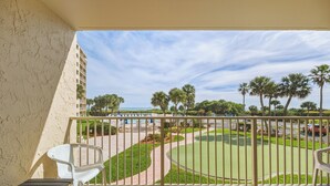 A balcony with a view of a golf course, palm trees, and a building in the background. - Oceanfront Balcony-Private Beach Access-Heated Pool (Cape Canaveral)