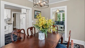 A dining room with a wooden table, chairs, and a vase of yellow flowers. There is a kitchen in the background with white cabinets and a refrigerator. - Blue Bungalow |  2BR 1BA | Lexington (Lexington)