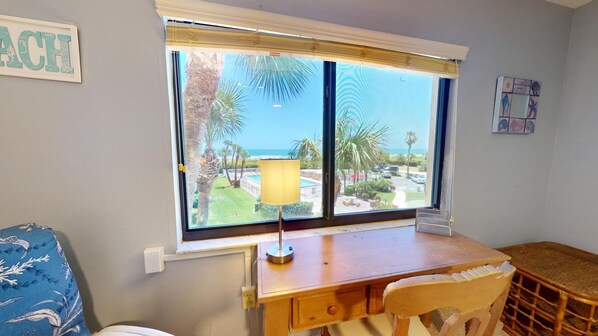 Ein Zimmer mit Schreibtisch aus Holz, einem Sessel, einer Lampe und Blick auf Pool und Palmen durch ein großes Fenster.