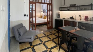 A compact room with a sofa, a dining table with chairs, a kitchen area with a sink and countertop, and a bed with a wooden frame. - Casa Lluavi (Mérida)