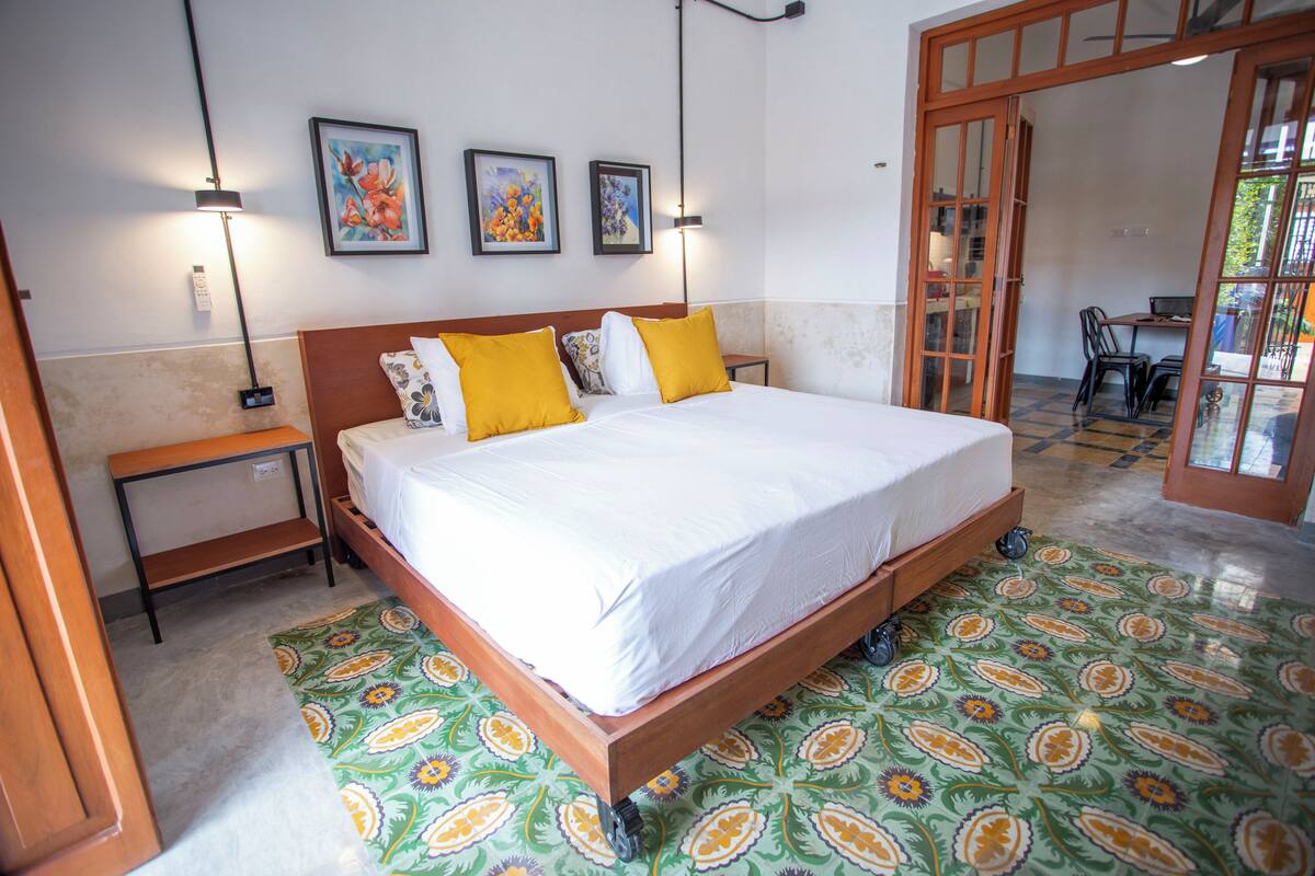 A bedroom with a wooden bed, white bedding, and a patterned floor. There are two bedside tables, a wall-mounted lamp, and framed artwork above the bed.