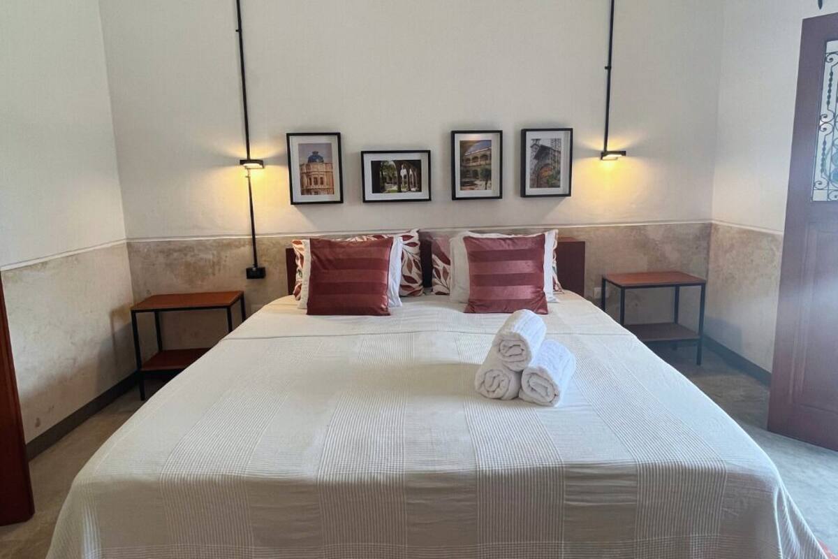 A neatly made bed with a white bedspread and two red pillows, a small table beside it, and a wall with framed pictures.
