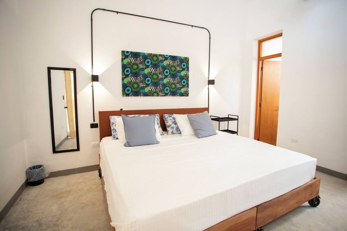 A neatly made bed with a wooden frame and wheels, a colorful abstract painting above it, a mirror on the wall, and a door to the right.