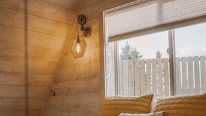 Une chambre avec un plafond en bois, un lit recouvert d’une courtepointe, une fenêtre avec des stores et un luminaire fixé au mur.