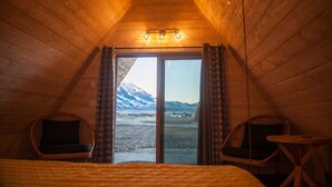 Ein gemütliches Zimmer mit Holzwänden und einer geneigten Decke, ausgestattet mit zwei Korbstühlen, einem kleinen Tisch und einem Bett. Ein großes Fenster bietet Blick auf eine verschneite Berglandschaft.
