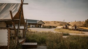 A rustic outdoor setting with a large tent-like structure, a covered seating area, and a dirt path leading to a hill.