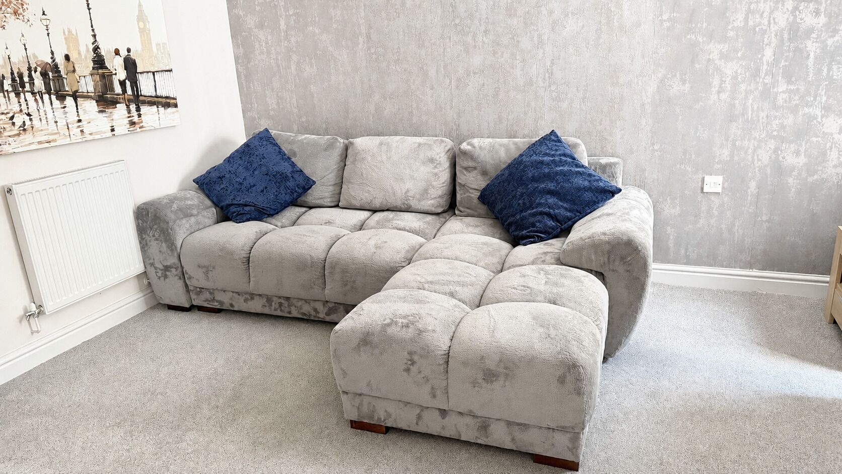 A grey sectional sofa with blue cushions, a radiator, and a textured grey wall.