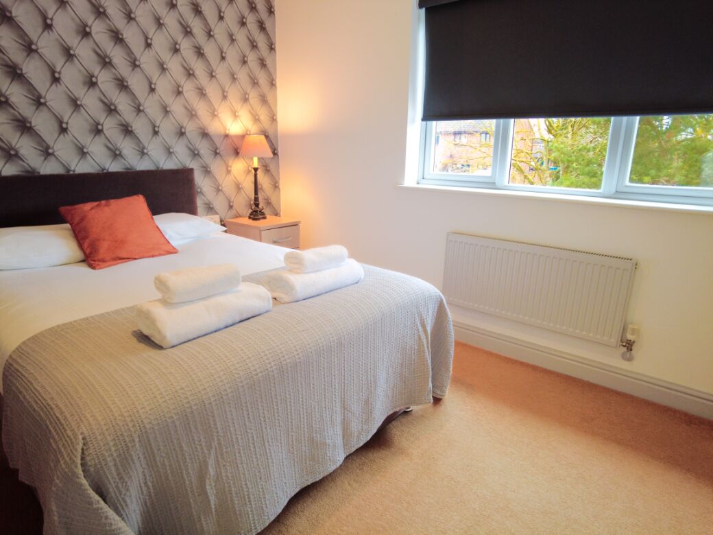 A neatly made bed with white linens and a grey blanket, a bedside lamp, a window with a view of greenery, and a radiator on the wall.