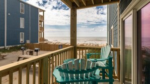 Een houten terras met stoelen met uitzicht op een strand, een gebouw met balkon, en een helder zicht op de oceaan.