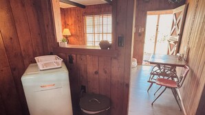 Een kleine keuken met een witte koelkast, een kleine prullenbak en een houten deur die leidt naar een eethoek met een tafel en stoelen.