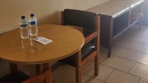Une petite table ronde en bois, sur laquelle sont posées deux bouteilles d’eau et un menu, un banc en bois avec un coussin, et un sol carrelé.