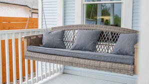 Eine Korbschaukel mit Kissen, befestigt an einem weißen Geländer, mit Blick auf eine Holzterrasse und ein Haus mit weißem Putz.