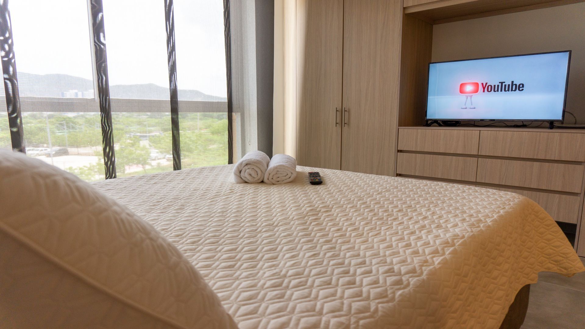 A hotel room with a bed, a TV displaying the YouTube logo, and a view of the outdoors.