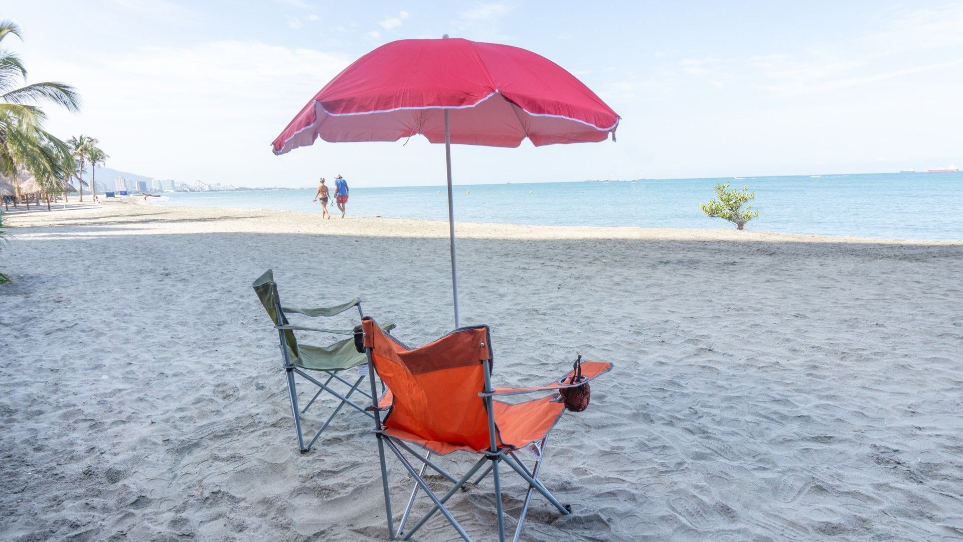 A beach with two folding chairs and a red umbrella.