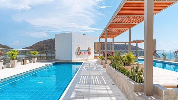A rooftop pool with a clear view of the sea and hills, a covered seating area, and potted plants.