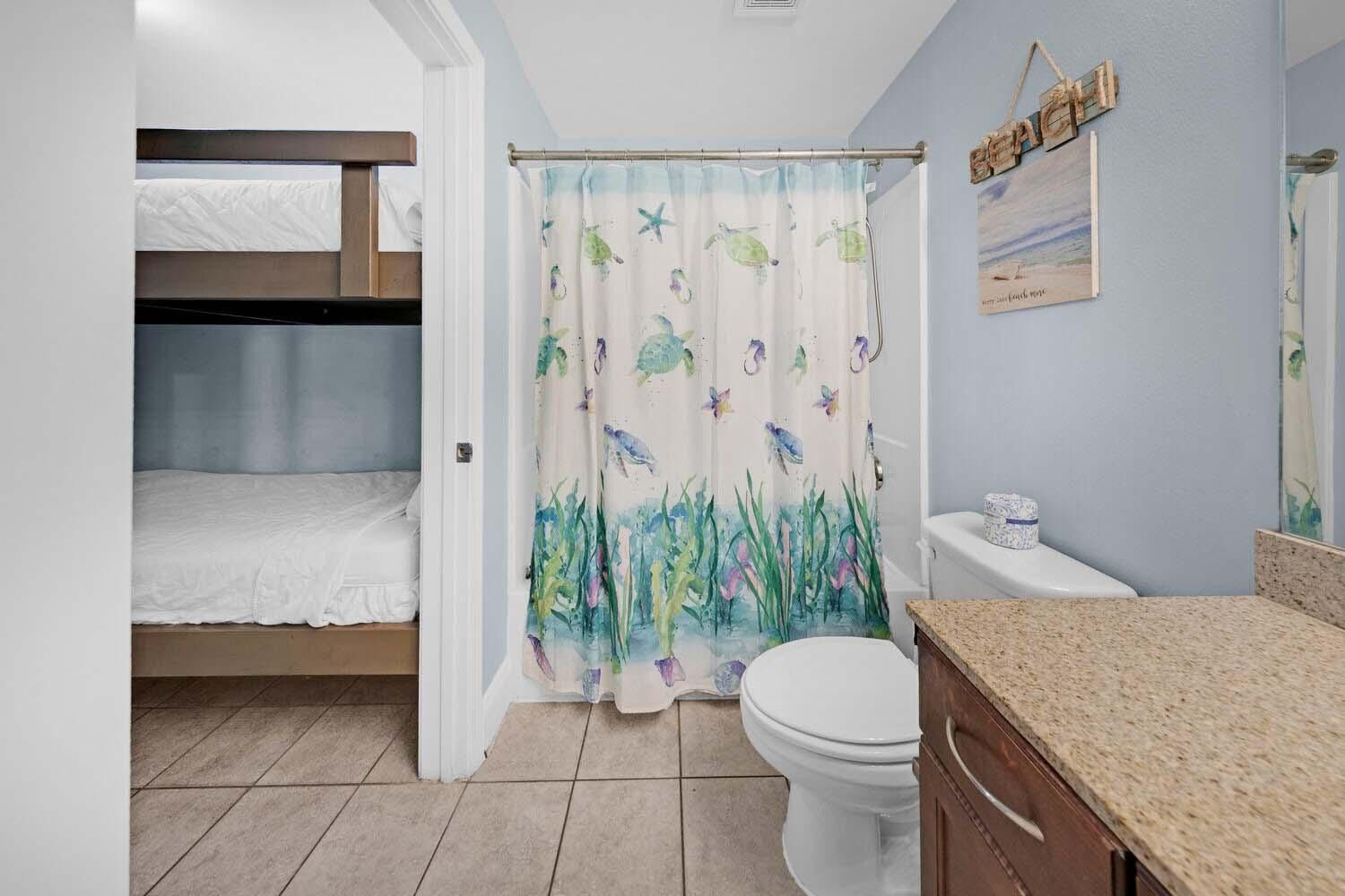 Een badkamer met een douchegordijn met aquatische motieven, een toilet, een wastafel met een granieten aanrechtblad, en een stapelbed in de aangrenzende kamer.